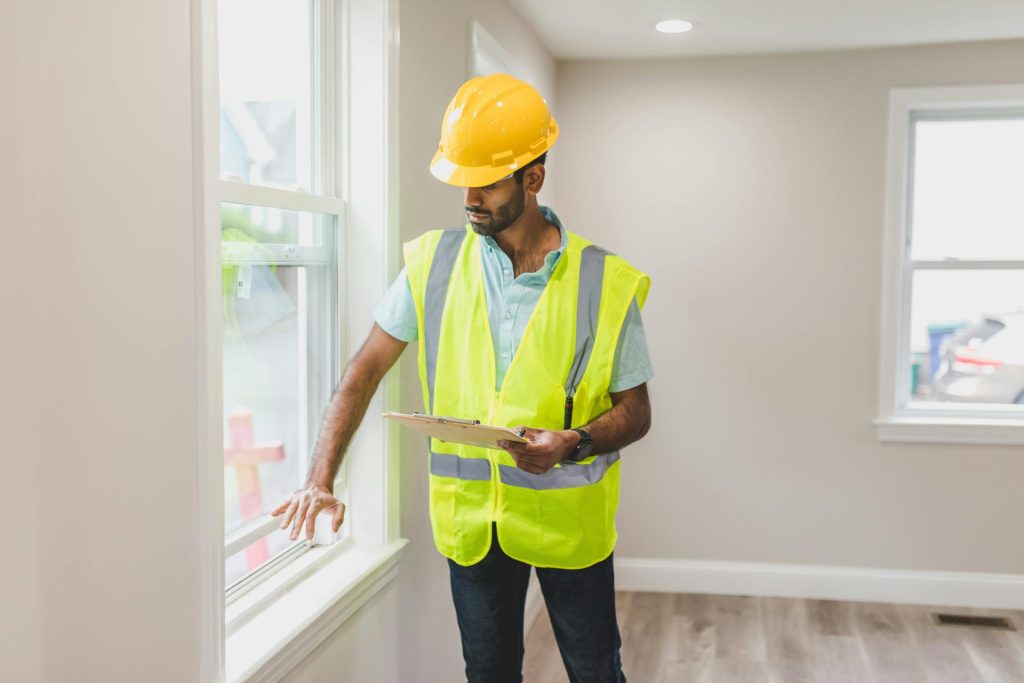 Ein Immobilienmakler inspiziert von innen ein Fenster; er trägt eine Sicherheitsweste und einen Schutzhelm, um die Sicherheit des Hauses zu gewährleisten.